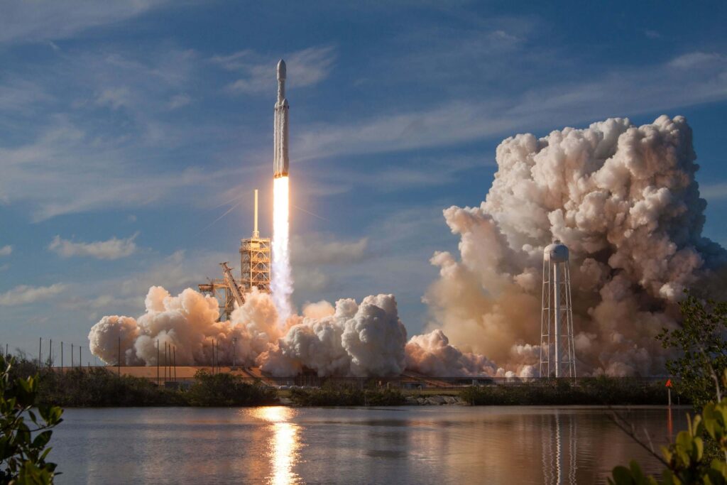 Rocket launching into a blue sky, trailing plumes of smoke, with a body of water in the foreground.